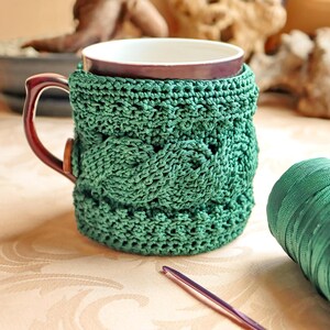May include: A handmade green crochet mug cozy wraps a brown mug. The cozy features a cable knit design. A ball of green yarn and a crochet hook are next to the mug. The mug has a white interior.