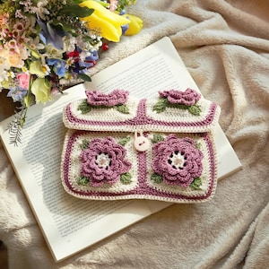 Pode incluir: Embreagem de crochê feita à mão com um design floral. A bolsa branca suja apresenta grandes flores roxas com centros de pérolas e folhas verdes. As bordas são aparadas com uma borda roxa. Um pequeno botão prende a aba.