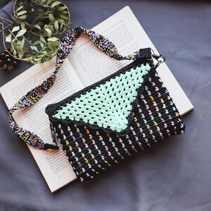 May include: A rectangular crochet crossbody bag featuring a black base with vertical multicolored yarn accents and a mint green triangular flap. The bag includes a detachable, patterned crochet strap with black, white, yellow, and pink threads.