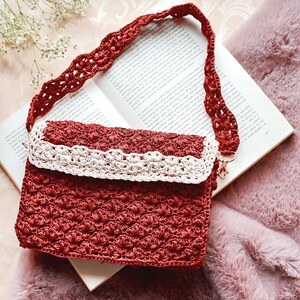 May include: A red crocheted handbag with a cream-colored trim and strap. The bag is rectangular with a flap closure and a long, decorative strap. The bag is resting on an open book.