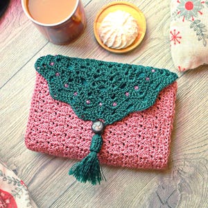 May include: A rectangular crochet pouch featuring a dusty rose body with a shell stitch pattern and a dark green triangular flap. The flap is adorned with small pink beads and a central dark button, finished with a matching green yarn tassel.