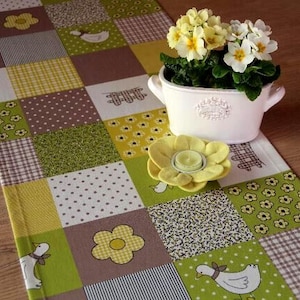 Könnte beinhalten: Ein grüner und brauner Patchwork-Tischläufer mit gelbem Blumendesign. Ein weißer Keramiktopf mit einem gelben Blumenarrangement befindet sich auf dem Tischläufer.