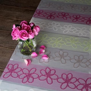 Könnte beinhalten: Ein Strauß rosa Rosen in einer klaren Glasvase auf einem Tischläufer mit Blumenmuster. Der Läufer hat ein sich wiederholendes Muster aus rosa, weißen und grünen Blumen.