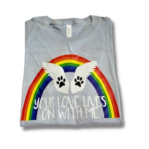 May include: Light blue t-shirt with a rainbow and two white wings with black paw prints. The text "Your love lives on with me" is printed on the shirt.