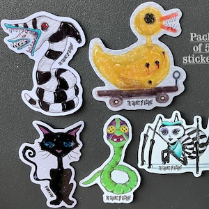 May include: A pack of five stickers featuring quirky, cartoonish creatures. The stickers include a black and white striped worm with a toothy grin, a yellow duck on a cart, a black cat, a green snake, and a spider. Text reads "Strange Creatures Coraline's Vibes".