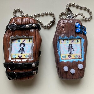 Puede incluir: Dos llaveros artesanales marrones en forma de barril con cadenas de bolas plateadas. Cada llavero presenta una pequeña pantalla que muestra un personaje de dibujos animados. Uno tiene correas negras y detalles plateados, el otro una mariposa azul.