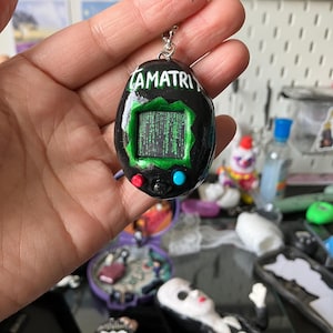 May include: A black pendant resembling a retro handheld game, with the word "AMATRIX" in white. The screen shows a green matrix-style code. It has red and blue buttons. A silver chain is attached to the top.