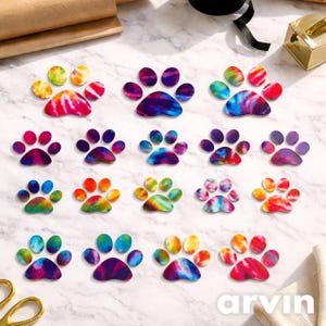 May include: A collection of colorful paw print stickers with tie-dye patterns. The stickers feature a variety of colors, including pink, purple, blue, green, and yellow. The paw prints are arranged on a white marble surface.