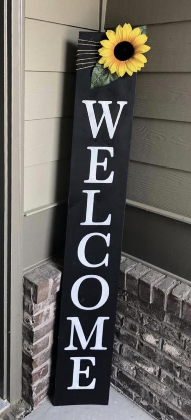 May include: A black wooden welcome sign with white letters. A yellow sunflower with green leaves is attached to the top of the sign.