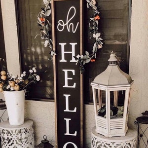 May include: A black wooden sign with white lettering that reads "oh hello" is leaning against a white door. The sign is decorated with a floral wreath. Two white lanterns are on either side of the sign.