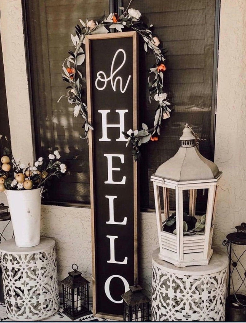 May include: A black wooden sign with white lettering that reads "oh hello" is leaning against a white door. The sign is decorated with a floral wreath. Two white lanterns are on either side of the sign.