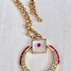 May include: Gold-toned necklace featuring a square pendant with a central red gem and a circular pendant with clear rectangular accents. The necklace has a beaded chain and a lobster clasp.