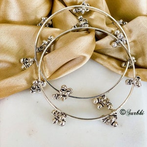 May include: Two silver bangles with decorative round accents. The bangles are stacked on top of each other and are on a gold fabric background.