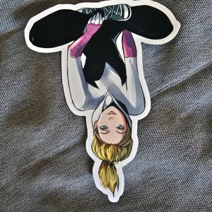 May include: A sticker of a female superhero wearing a white and black suit with pink spider webbing. She is sitting with her legs crossed and her head upside down. She has blonde hair in a ponytail and green eyes.