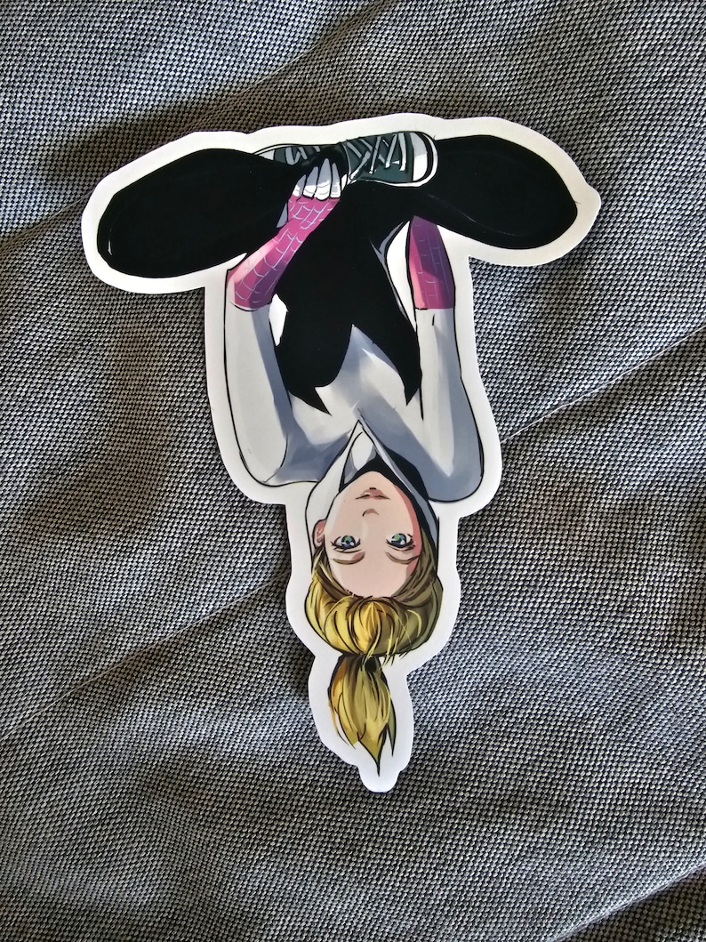 May include: A sticker of a female superhero wearing a white and black suit with pink spider webbing. She is sitting with her legs crossed and her head upside down. She has blonde hair in a ponytail and green eyes.