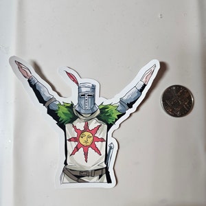 May include: A white sticker with a cartoon illustration of a knight wearing a helmet and armor, holding his arms up in the air. The knight is wearing a white tunic with a red sun and a face on it. The knight is standing on a green patch of grass.