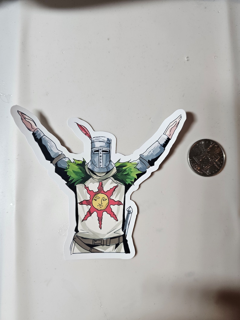 May include: A white sticker with a cartoon illustration of a knight wearing a helmet and armor, holding his arms up in the air. The knight is wearing a white tunic with a red sun and a face on it. The knight is standing on a green patch of grass.