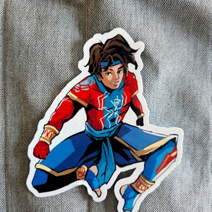 May include: A sticker of a cartoon character wearing a red and blue outfit with a gold belt. The character has brown hair and is in a jumping pose.