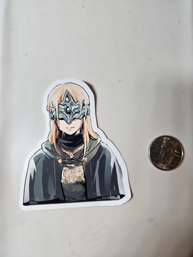 May include: A sticker featuring a character from a video game. The character is a woman with long blonde hair, wearing a dark cloak and a silver mask with a large, central gem. The sticker is on a white background.