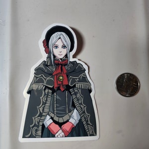 May include: A sticker featuring a cartoon illustration of a woman with white hair, wearing a black hat, a black cloak, and a red bow. She has blue eyes and is holding her hands together in front of her.