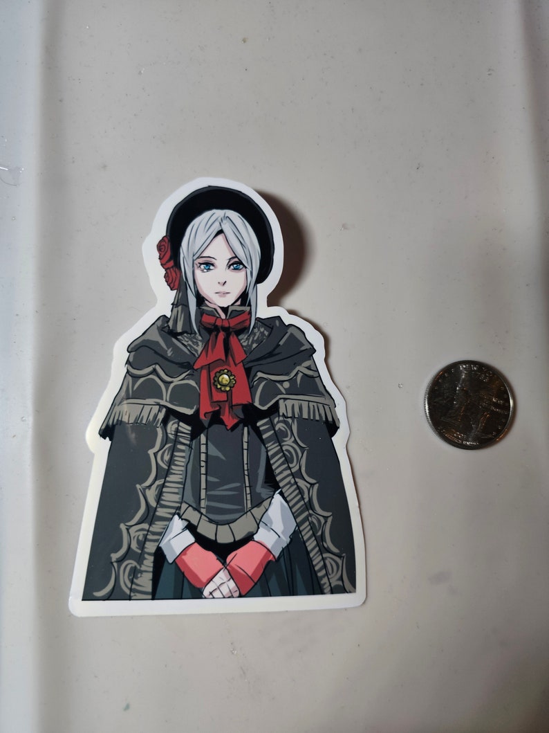 May include: A sticker featuring a cartoon illustration of a woman with white hair, wearing a black hat, a black cloak, and a red bow. She has blue eyes and is holding her hands together in front of her.