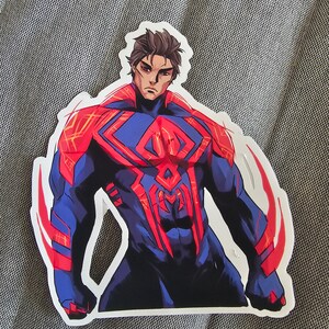 May include: A sticker featuring a superhero in a red and blue suit with a spider symbol on the chest. The superhero has a determined expression and is posed in a heroic stance.