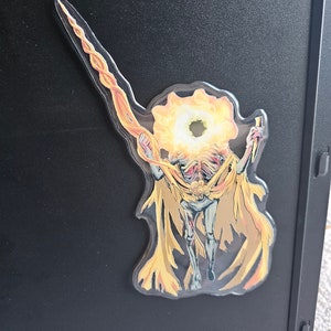 May include: A clear sticker featuring a stylized illustration of a figure with a fiery halo and cape. The figure is depicted in shades of yellow, orange, and gold, with a skeletal appearance. The sticker has a long, flame-like extension.