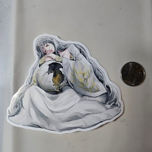 May include: A sticker featuring a woman with long white hair, wearing a white robe with gold accents, holding a large egg in her arms. The woman has a sad expression on her face.