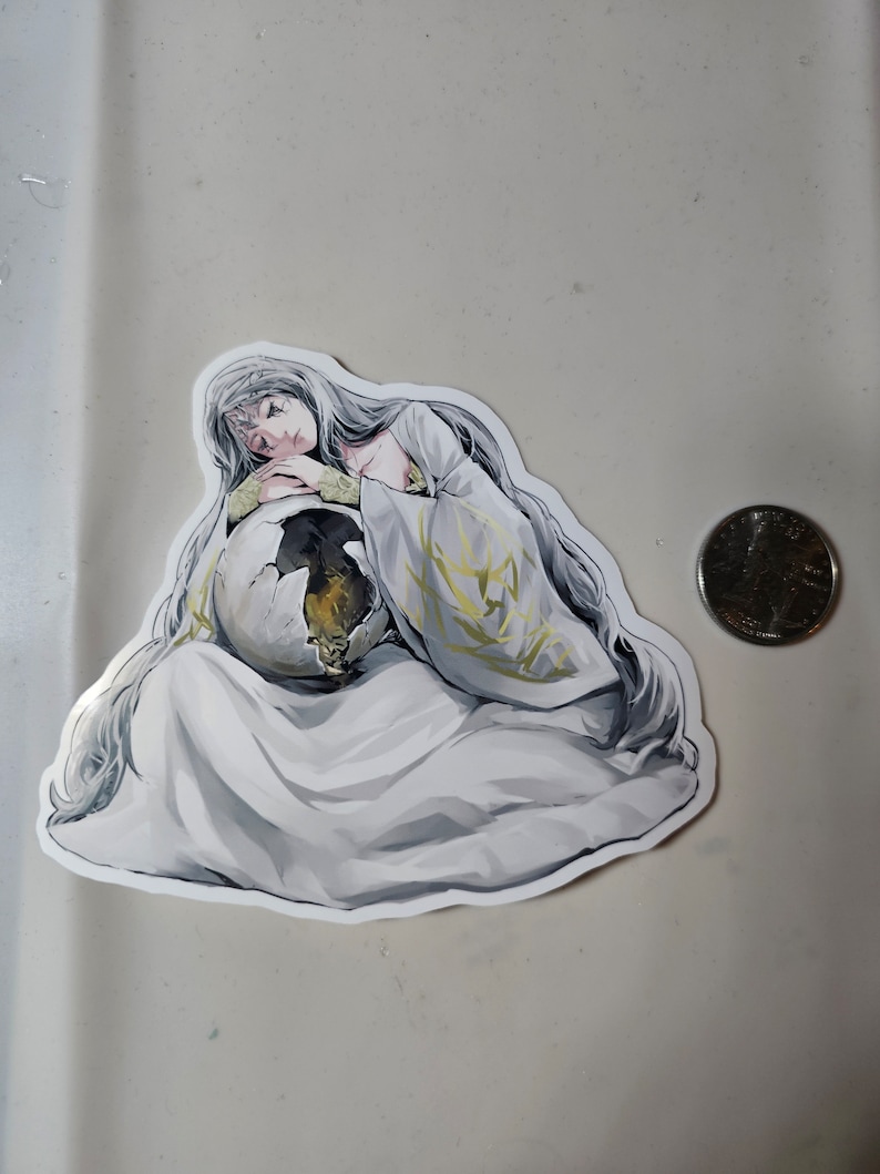 May include: A sticker featuring a woman with long white hair, wearing a white robe with gold accents, holding a large egg in her arms. The woman has a sad expression on her face.