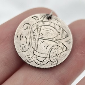 May include: A small, round, silver-toned pendant with an engraved design. The pendant features an ornate letter "R" in the center, surrounded by decorative lines and patterns. A small loop is at the top for attaching to a chain or bracelet.