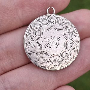 May include: A circular silver-toned pendant featuring an intricate engraved star pattern surrounding a central monogram with the letters HC in cursive script. The pendant includes a small metal loop at the top for attachment to a chain or cord.