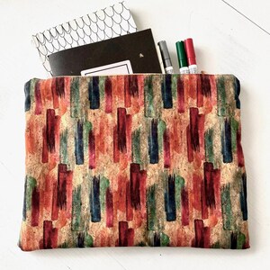 Könnte beinhalten: Eine Reißverschlusstasche aus Korkstoff mit einem farbenfrohen abstrakten Muster aus roten, orangen, blauen und grünen Pinselstrichen. Die Tasche liegt flach und offen und zeigt ein Notizbuch und Stifte im Inneren.