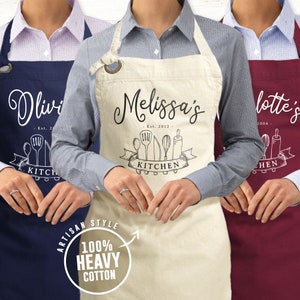 May include: Three personalized aprons with a kitchen theme. The aprons are navy blue, beige, and burgundy. Each apron has a name, established date, and a graphic of kitchen utensils. The text on the aprons reads "Olivia's Est. 20", "Melissa's Est. 2012", and "Lotte's 2004". The aprons are made of 100% heavy cotton.
