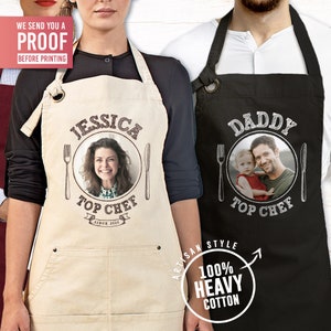May include: Two personalized aprons with a photo, text, and a fork and knife design. The aprons are beige and black with the text "Jessica Top Chef Since 2023" and "Daddy Top Chef". The aprons are made of 100% heavy cotton.