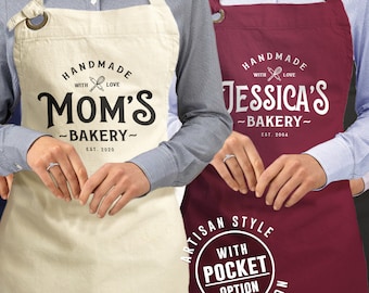 Könnte beinhalten: Zwei weiße Baumwollschürzen mit schwarzem Text. Die Schürze links sagt "Handmade with love Mom's Bakery Est. 2020". Die Schürze rechts sagt "Handmade with love Jessica's Bakery Est. 2004". Beide Schürzen haben einen Kreis unten, der "Artisan Style with Pocket Option Heavy Cotton" sagt.