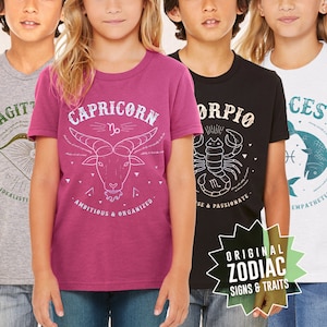 May include: Four children's t-shirts with zodiac signs and traits. The shirts are pink, black, gray, and white. The signs are Capricorn, Scorpio, Sagittarius, and Pisces.