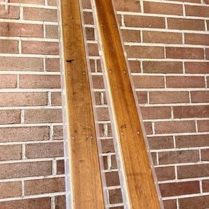 May include: Two wooden boards with a clear plastic edge, secured with small silver screws. The boards are a warm brown color and are leaning against a red brick wall. The boards are likely for a construction project.