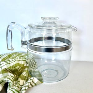 May include: A clear glass coffee pot with a lid and handle, featuring a silver-coloured metal band around the middle. A green and white chequered cloth is draped on the side. The pot is designed for brewing coffee.