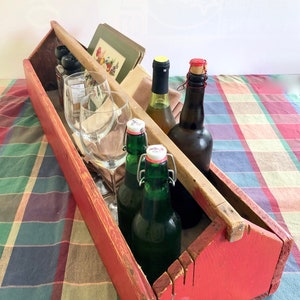 May include: A red wooden tool box with a handle, filled with glass bottles and wine glasses. The box is sitting on a checked tablecloth.