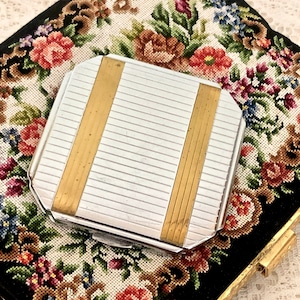 May include: A silver and gold compact mirror with a floral design on the background. The mirror has a rectangular shape with rounded corners and a gold trim.