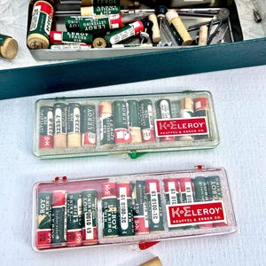 May include: An open green metal case with lettering tools, including pens and nibs. Below are two clear plastic cases filled with lettering pens, labeled "LEROY" and "K+E LEROY". A wooden cylinder is at the bottom.