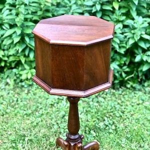 Può includere: Un tavolino vintage in legno marrone scuro con piano e corpo ottagonali. Il tavolo ha una base a piedistallo con un design a quattro gambe. Il tavolo è appoggiato su una superficie erbosa, con fogliame verde sullo sfondo.