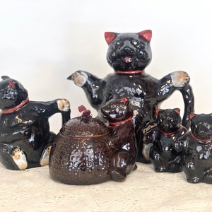 May include: A black ceramic teapot and sugar bowl shaped like bears, with three smaller bear-shaped creamer and salt and pepper shakers. The teapot has a red rim and a red collar around its neck. The sugar bowl has a brown and black honeycomb pattern. The smaller bears have red collars around their necks.