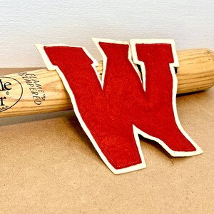 Può includere: Una mazza da baseball Louisville Slugger in legno con una lettera "W" in feltro rosso con bordo bianco. La mazza è marrone chiaro con testo nero. La lettera è rossa con bordo bianco.