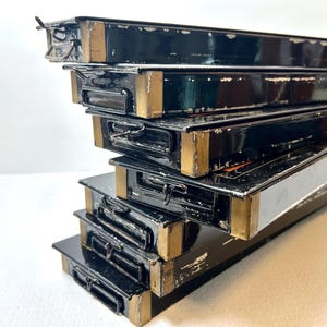 May include: A stack of rectangular black metal trays with gold-toned metal corner accents and small black metal handles on the front edges. The black finish shows signs of wear and paint loss along the edges, revealing the underlying metal surface.