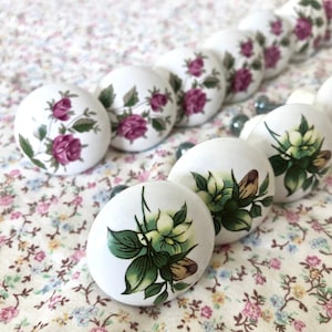 May include: A collection of white ceramic drawer knobs with floral designs. The knobs feature pink roses, green leaves, and white flowers.