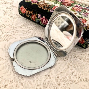 May include: A silver compact mirror with a round mirror on one side and a round, empty space on the other side. The compact is open and the mirror is facing up.