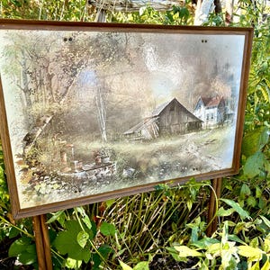 May include: A framed landscape painting showing a rural scene with a barn, house, and trees. The artwork has a warm colour palette with brown, beige, and green tones. The painting is displayed on a wooden easel outdoors.