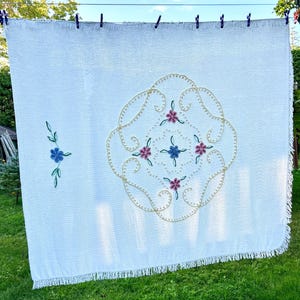 May include: White embroidered blanket with a floral design. The blanket has a large central embroidered pattern with pink, blue, and gold accents. A smaller floral design is on the left side. The blanket is hanging on a washing line.
