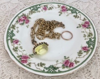 May include: A white porcelain trinket dish with a floral pattern featuring pink roses and green leaves. A gold chain necklace with a yellow glass pendant is resting on the dish.